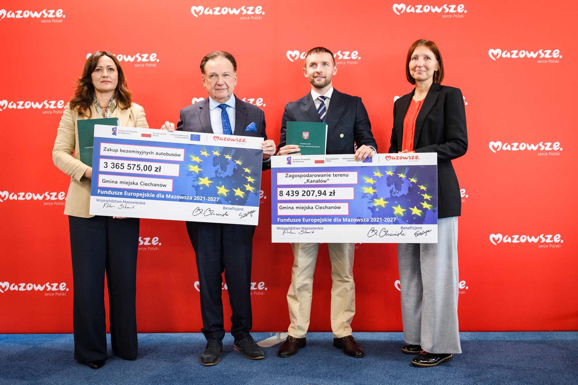 Cztery osoby stoją w rzędzie na niebieskim dywanie przed czerwonym panelem z powtarzającym się białym logo „Mazowsze”, dwie środkowe osoby trzymają duże symboliczne czeki z motywem gwiazd UE i widocznymi kwotami 3 365 575,00 zł oraz 8 439 207,94 zł, a dwie kobiety po bokach trzymają teczki i dokumenty.